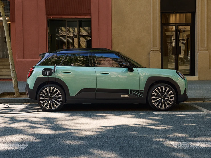 Zijaanzicht van de MINI Aceman Electric in Ocean Wave Green met Black Roof die langs een straat wordt opgeladen.