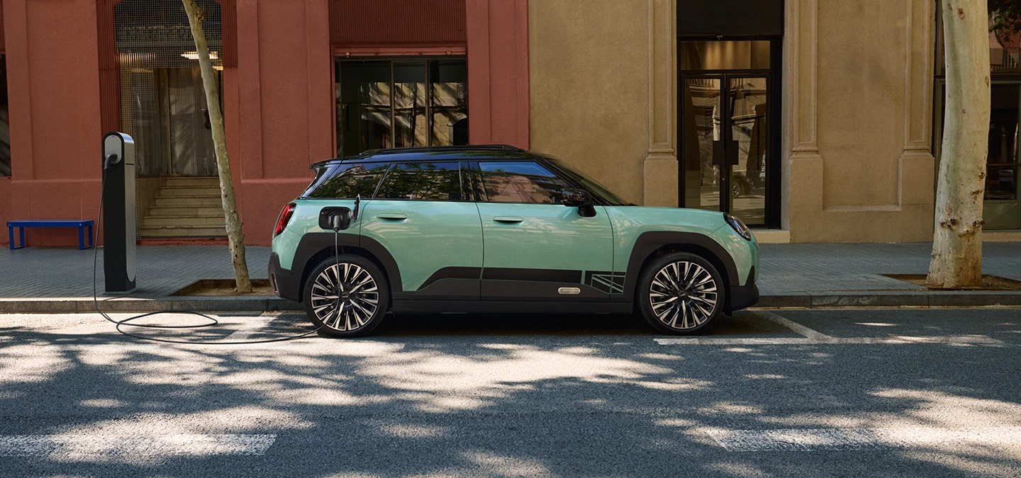 Zijaanzicht van de MINI Aceman Electric in Ocean Wave Green met Black Roof die langs een straat wordt opgeladen.