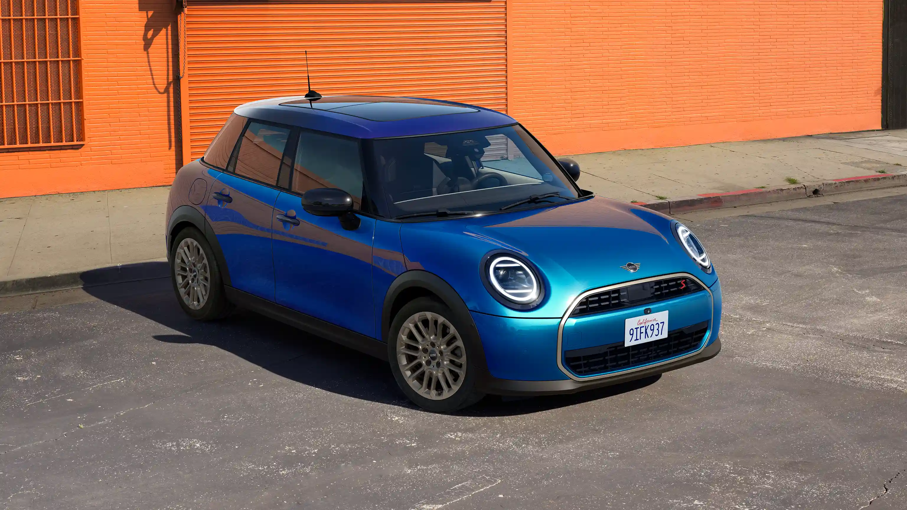 Vooraanzicht van een geparkeerde MINI Cooper 5-deurs in Icy Sunshine Blue met een Multitone Blue dak op straat met een oranje muur op de achtergrond