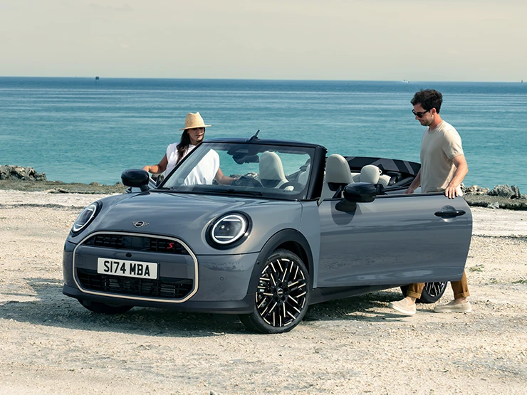 Vue avant gauche de la MINI Cooper Cabrio Cooper Grey au bord d’une plage avec deux personnes à côté.