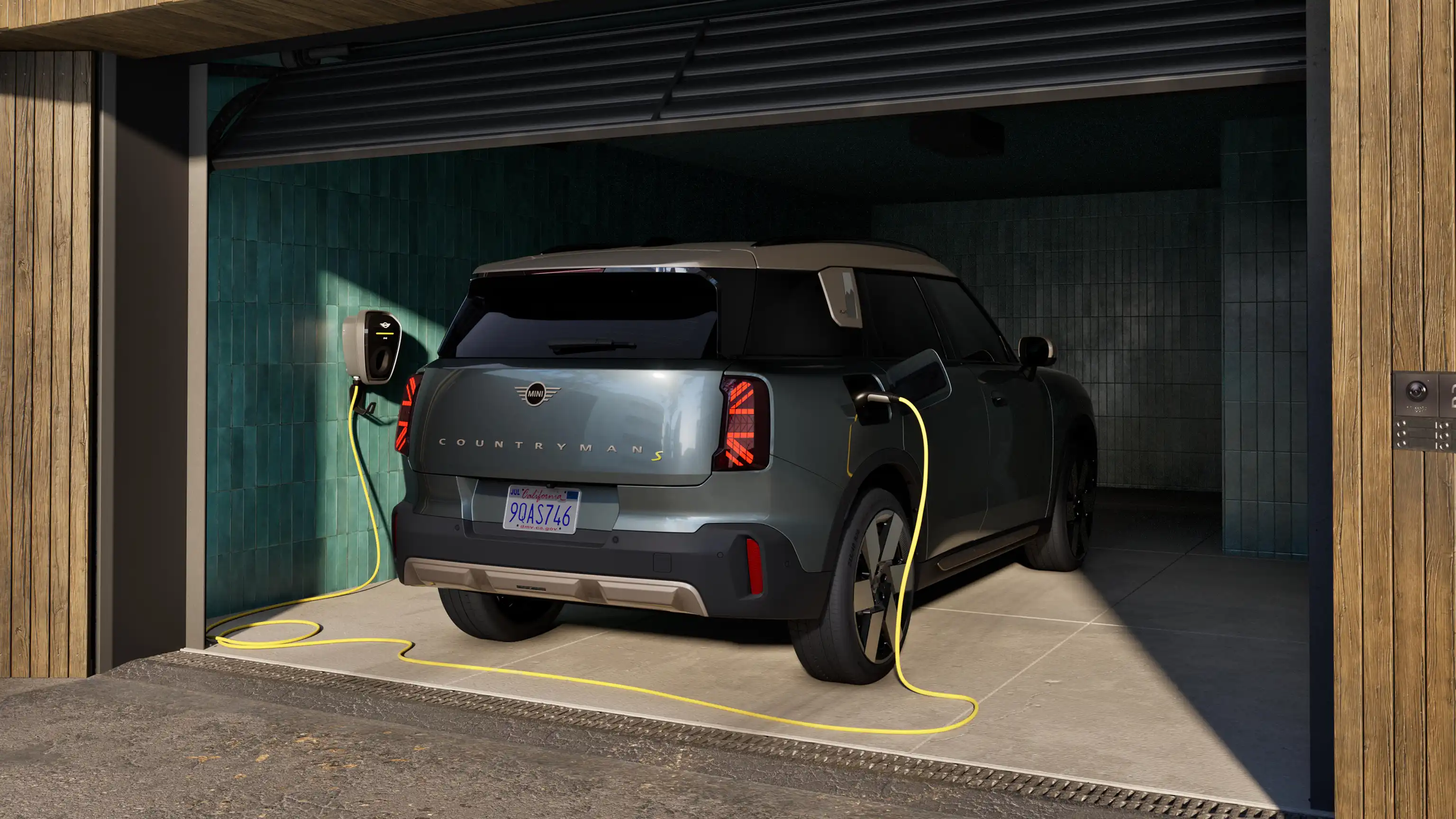 Vue arrière d’une MINI Countryman 100 % électrique en Smokey Green, stationnée dans un garage ouvert et reliée à un câble de recharge.