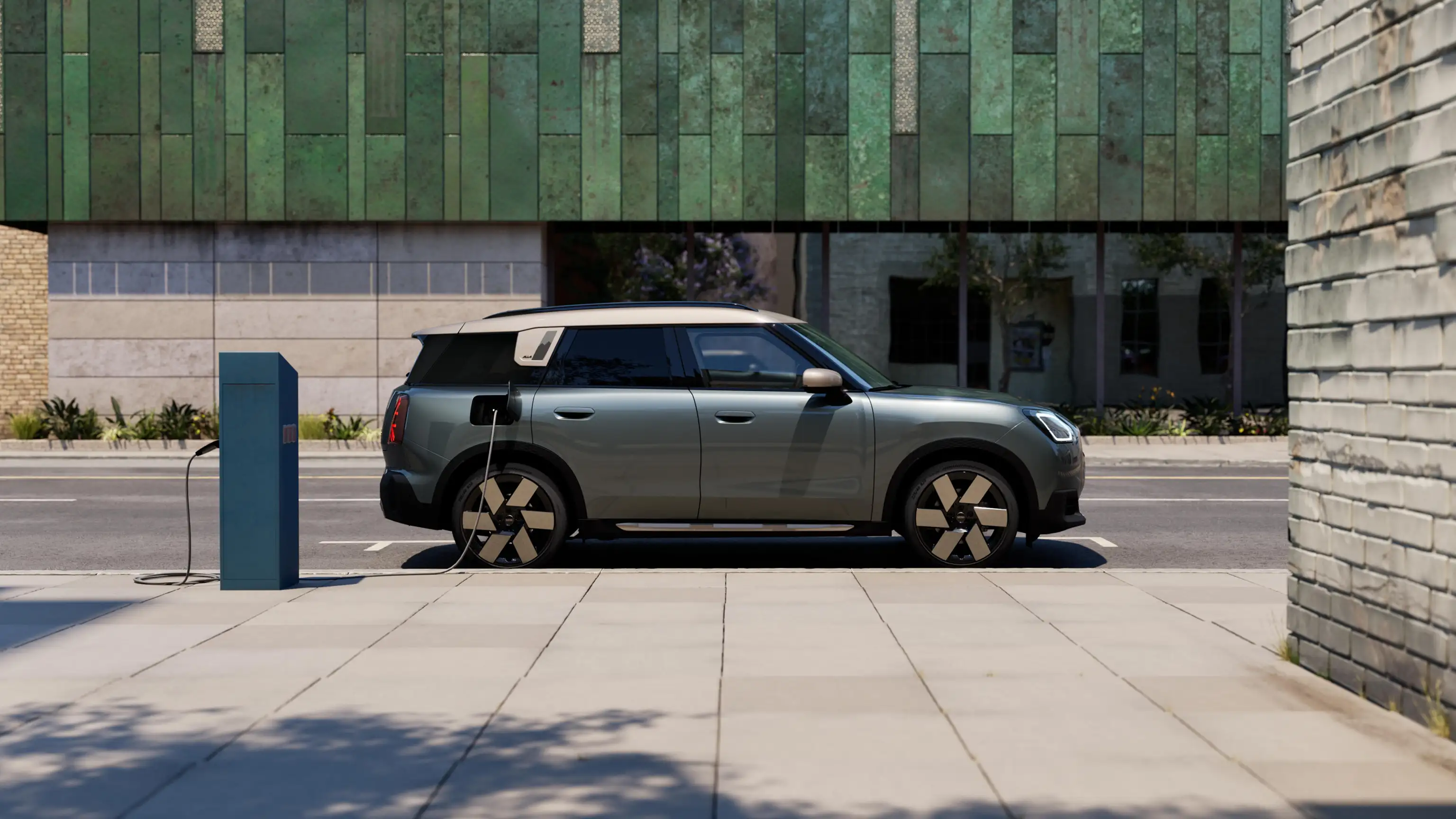 Vue latérale d’une MINI Countryman 100 % électrique en Smokey Green avec toit Vibrant Silver, en cours de recharge dans la rue