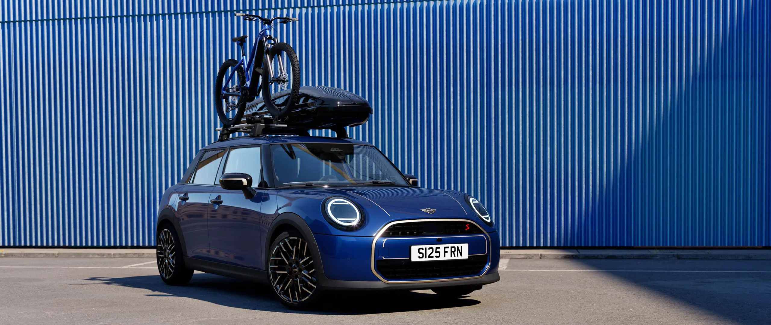 Vue de face de la MINI Aceman en Indigo Sunset Blue avec porte-vélos de toit MINI, transportant un vélo de montagne et un coffre de toit MINI 320.
