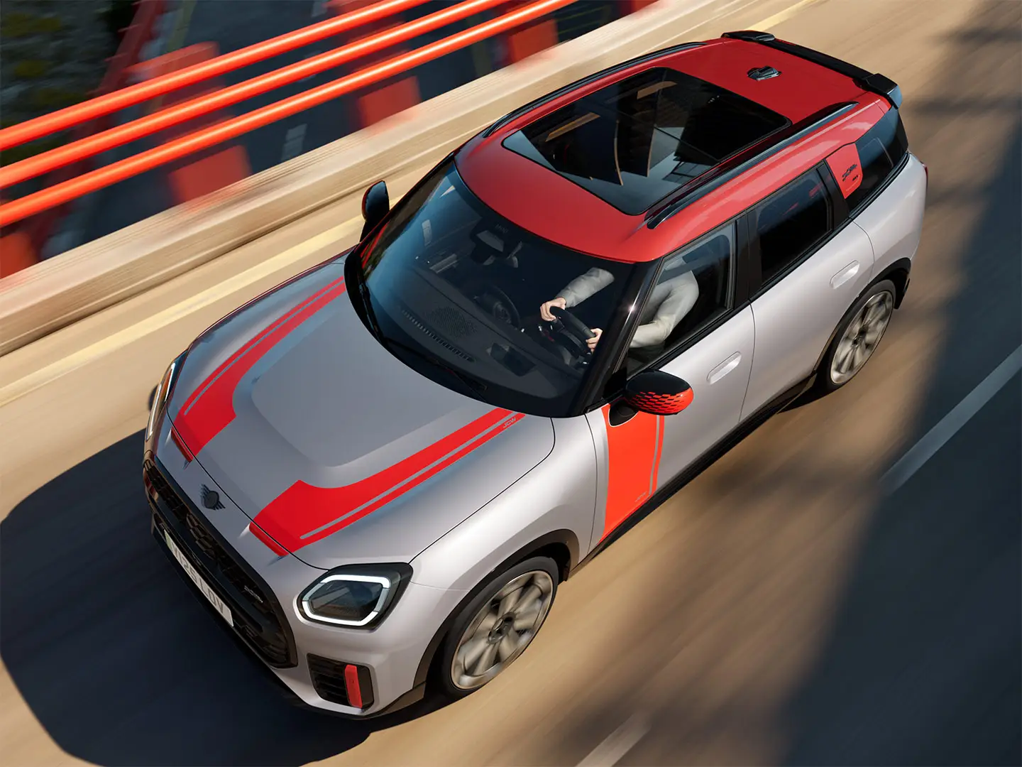 MINI Countryman John Cooper Works en teinte Melting Silver avec coques de rétroviseurs, bandes latérales et de capot, toit et ailettes en Chili Red, circulant en ville – vue de trois-quarts avant.