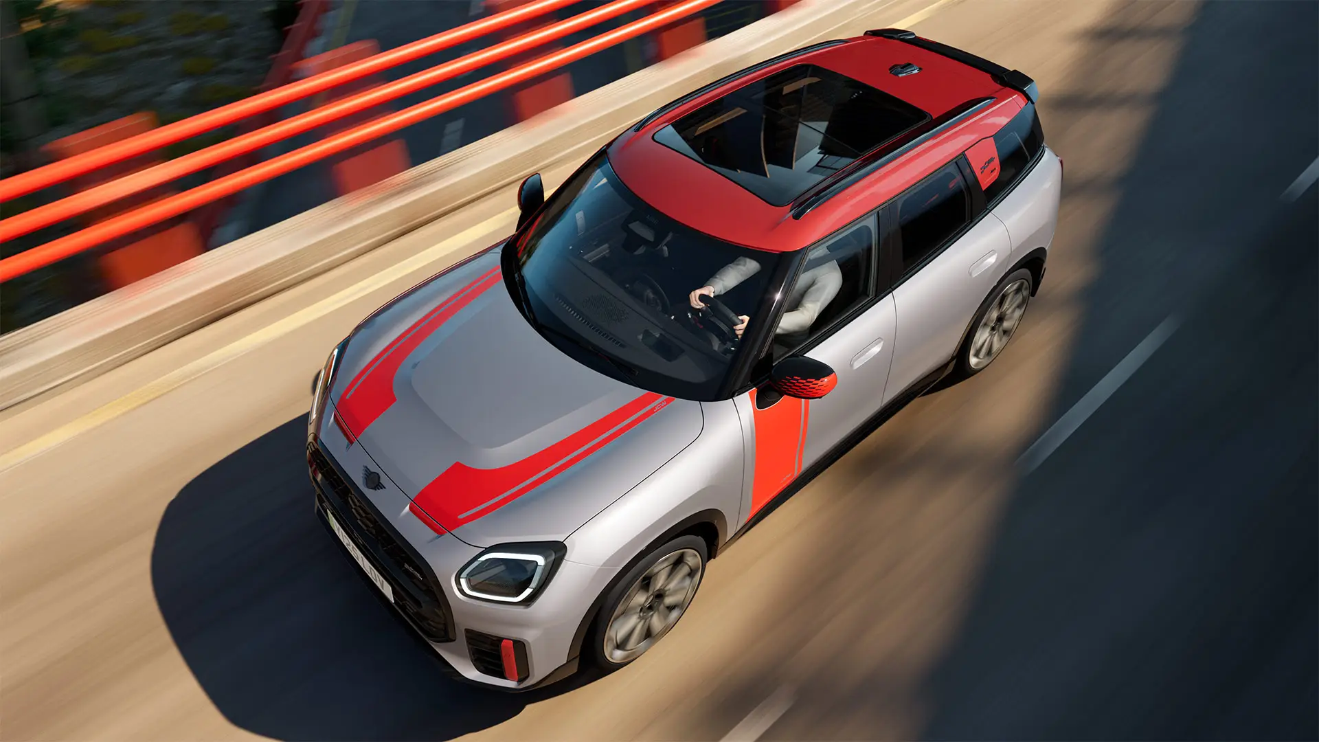 MINI Countryman John Cooper Works en teinte Melting Silver avec coques de rétroviseurs, bandes latérales et de capot, toit et ailettes en Chili Red, circulant en ville – vue de trois-quarts avant.