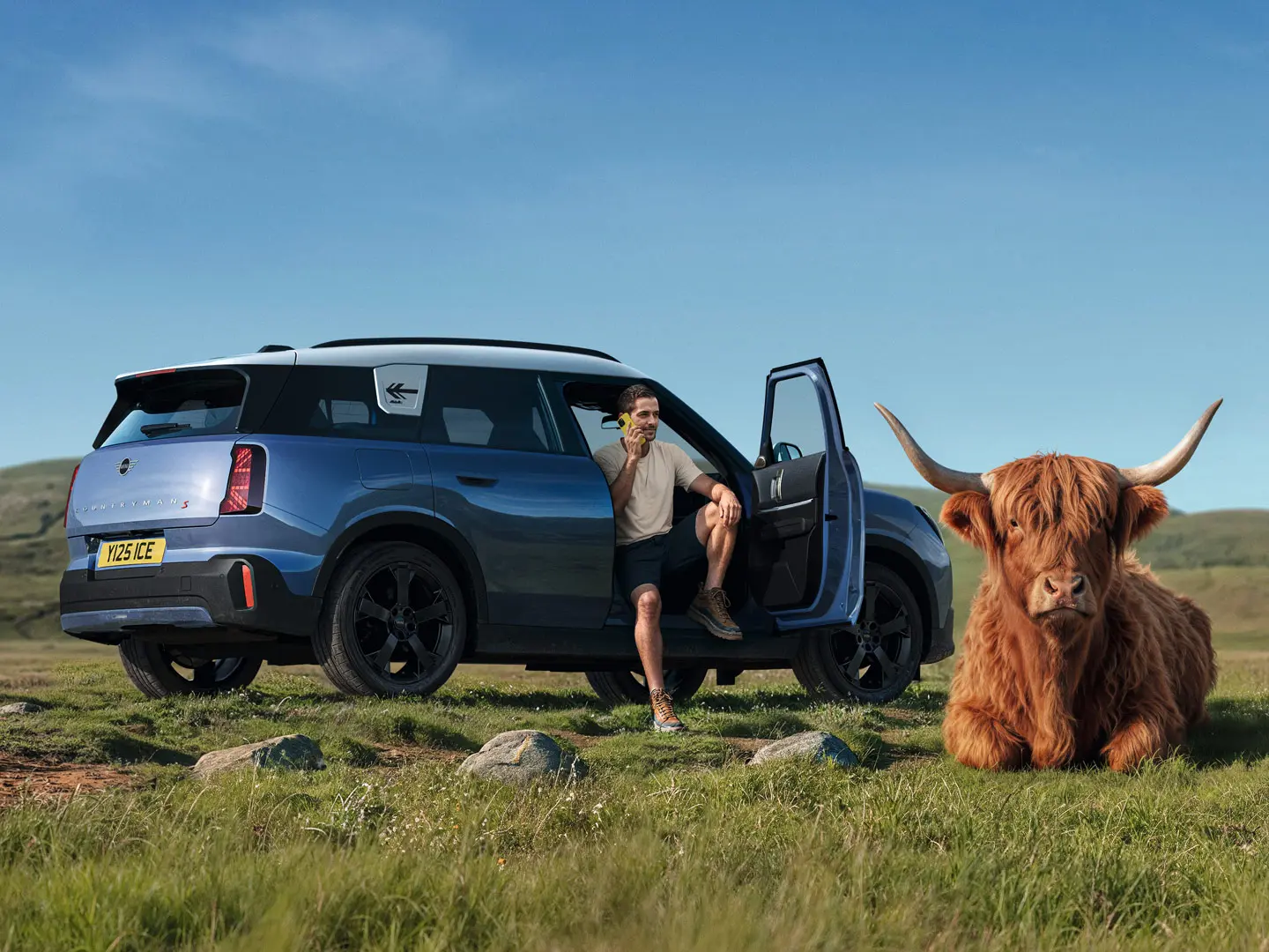 Un homme à l’extérieur de sa MINI Countryman S, en train de parler au service MINI Roadside Assistance via la MINI App, avec un taureau Highland couché à proximité.